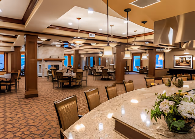 Spacious senior living dining room with a curved granite countertop in the foreground, multiple tables and chairs, pendant lights, and a fireplace in the background.