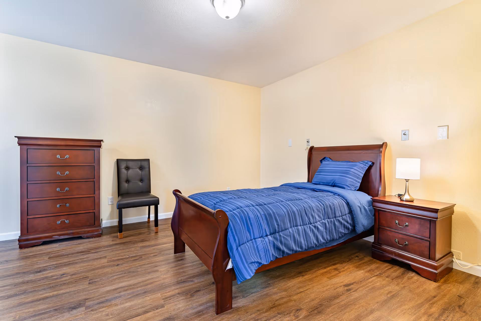 A simple bedroom with wooden flooring featuring a single wooden bed with blue striped bedding, a matching wooden nightstand with a lamp, a wooden chest of drawers, and a black cushioned chair against a light yellow wall.