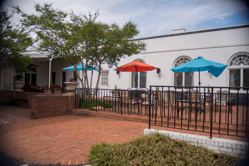 Outdoor patio area with brick flooring and black metal railings. There are tables with colorful umbrellas in red and blue, surrounded by chairs. The patio is adjacent to a white building with arched windows and some greenery including trees and bushes.