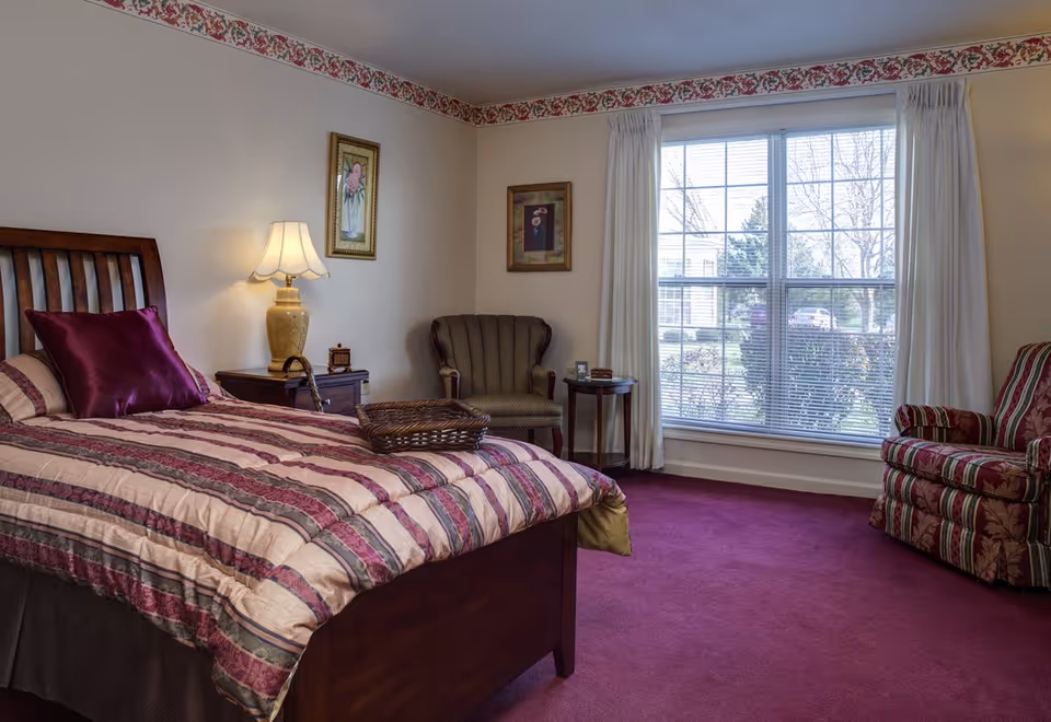 Furnished bedroom with a wooden bed dressed in striped bedding, two armchairs, a nightstand with a lamp, and a large window.