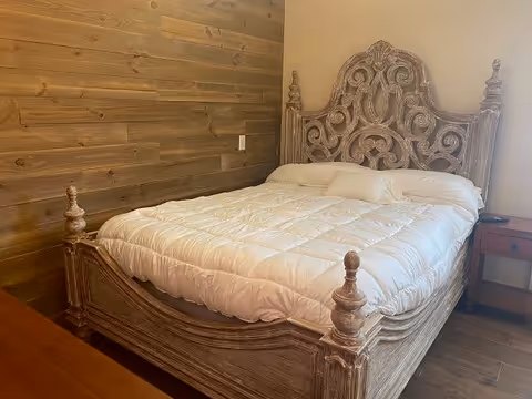 A bedroom with a wooden bed frame featuring intricate carvings and a white comforter. The room has a wooden accent wall and a small wooden nightstand with a drawer next to the bed.
