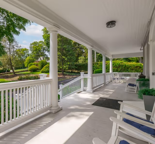 A spacious covered porch with white wooden railings and columns overlooking a green garden area. The porch has several white rocking chairs with blue cushions and a black doormat near the entrance. The scene is bright and sunny with trees and bushes visible in the background.