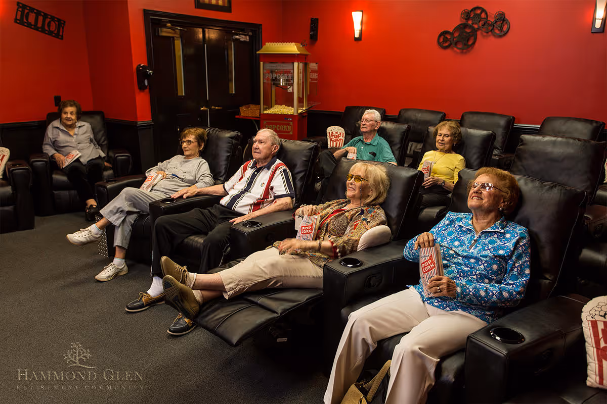 A group of elderly people sitting in black leather reclining chairs in a small movie theater room with red walls, watching a movie and holding popcorn.