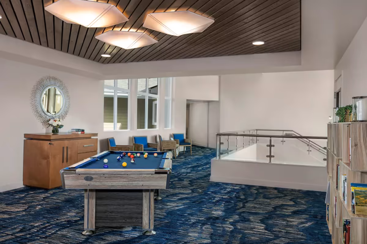 Interior common area with a pool table in the center, blue patterned carpet, wooden ceiling with modern light fixtures, a wooden cabinet with a decorative mirror and flowers, several chairs along the wall, and a bookshelf filled with books on the right side.