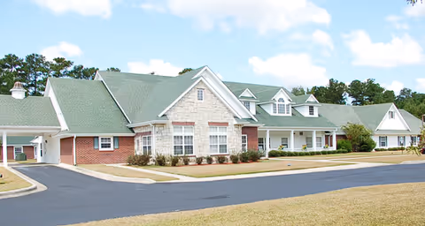 Exterior view of a single-story senior living facility building with a green roof, brick and stone facade, multiple windows, and a covered entrance. The building is surrounded by a paved driveway and landscaped grass areas under a partly cloudy sky.