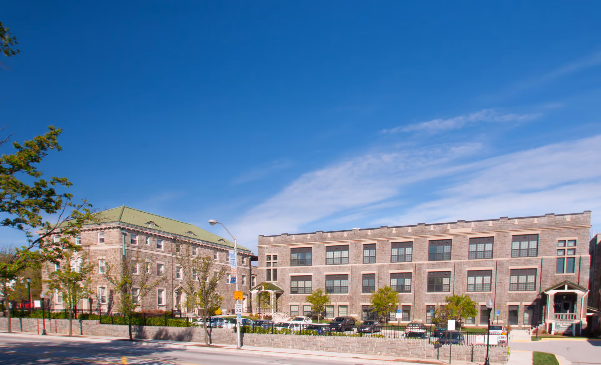 Exterior view of the John Manley House facility showing two large stone buildings with multiple windows under a clear blue sky. There is a parking area with several cars in front of the buildings, and a stone wall with a sidewalk and street in the foreground.