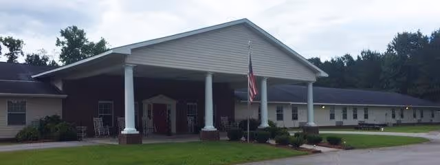 Front exterior view of The Gardens of Pamlico facility with a covered entrance supported by white columns, an American flag on a flagpole, and a well-maintained lawn and shrubs in front.