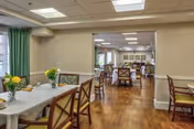 Interior view of a senior living facility dining room with wooden floors, tables covered with white tablecloths, chairs with cushions, and small flower arrangements on the tables. The room is well-lit with ceiling lights and has green curtains on the windows.