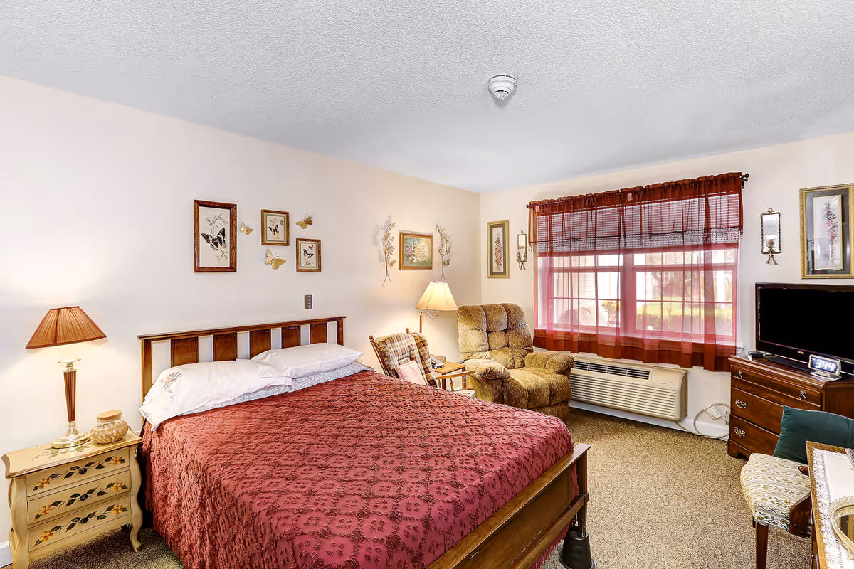 A cozy bedroom with a wooden bed covered in a red patterned bedspread, a nightstand with a lamp and decorative items, a comfortable armchair and rocking chair near a window with red curtains, a dresser with a TV and digital clock, and framed artwork on the walls.