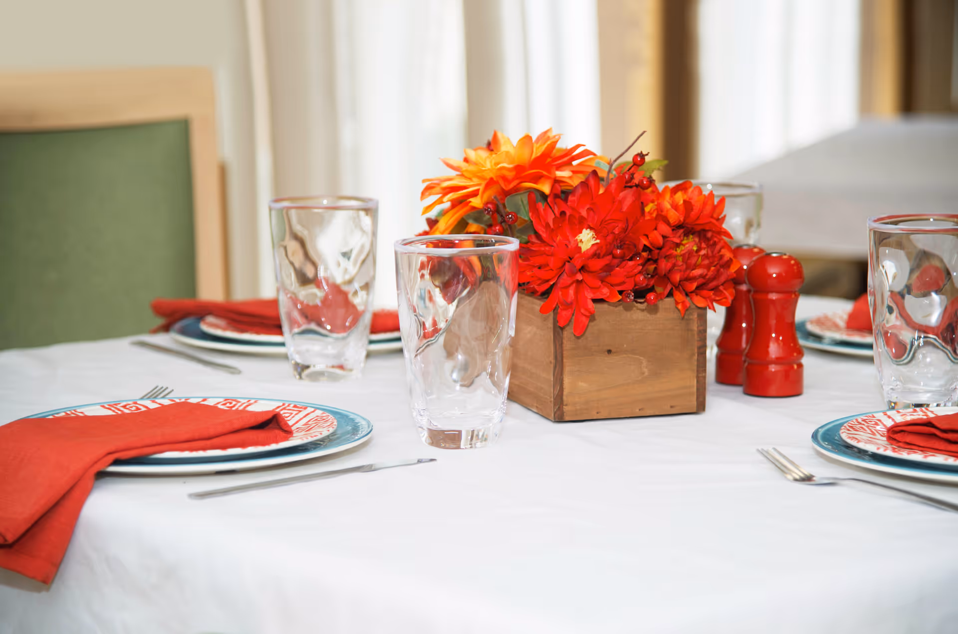 A dining table set with plates, red cloth napkins, clear drinking glasses, silverware, and a wooden box centerpiece filled with bright red and orange flowers. There are red salt and pepper shakers on the table, and a green chair is partially visible in the background.