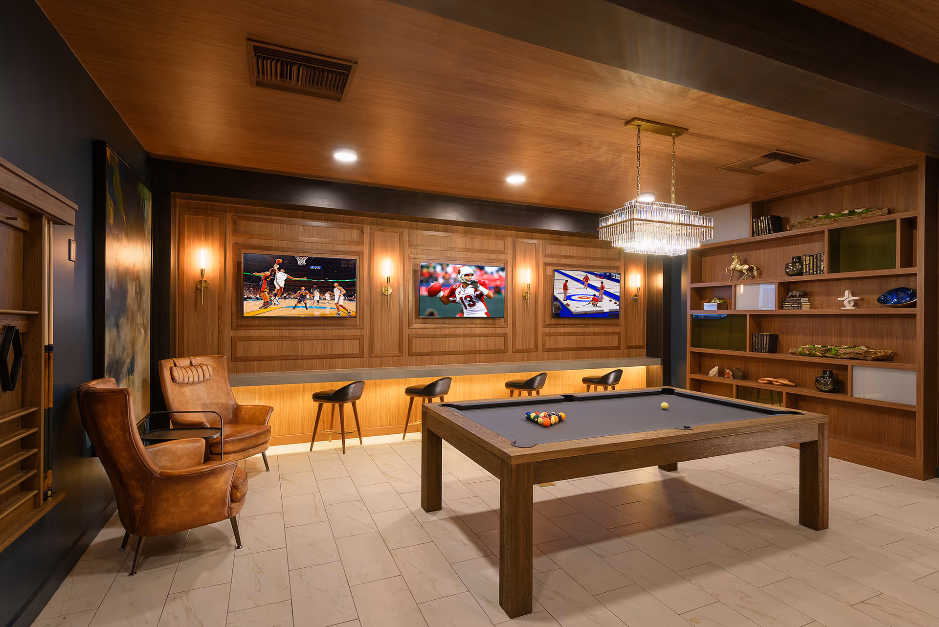 A stylish game room with a pool table in the center, surrounded by wooden walls and ceiling. There are four bar stools along a counter beneath three wall-mounted TVs showing different sports. Two brown leather armchairs are positioned near the left wall, and a large wooden bookshelf with decorative items is on the right. A modern chandelier hangs above the pool table.