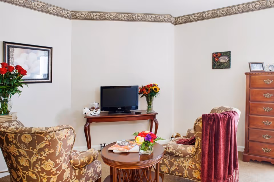 A cozy living room with two patterned armchairs, one draped with a red throw blanket, facing a small wooden table with a flower arrangement and magazines. A wooden console table holds a flat-screen TV and a vase of flowers. There is a wooden chest of drawers against the wall, and framed artwork and decorative items on the walls.
