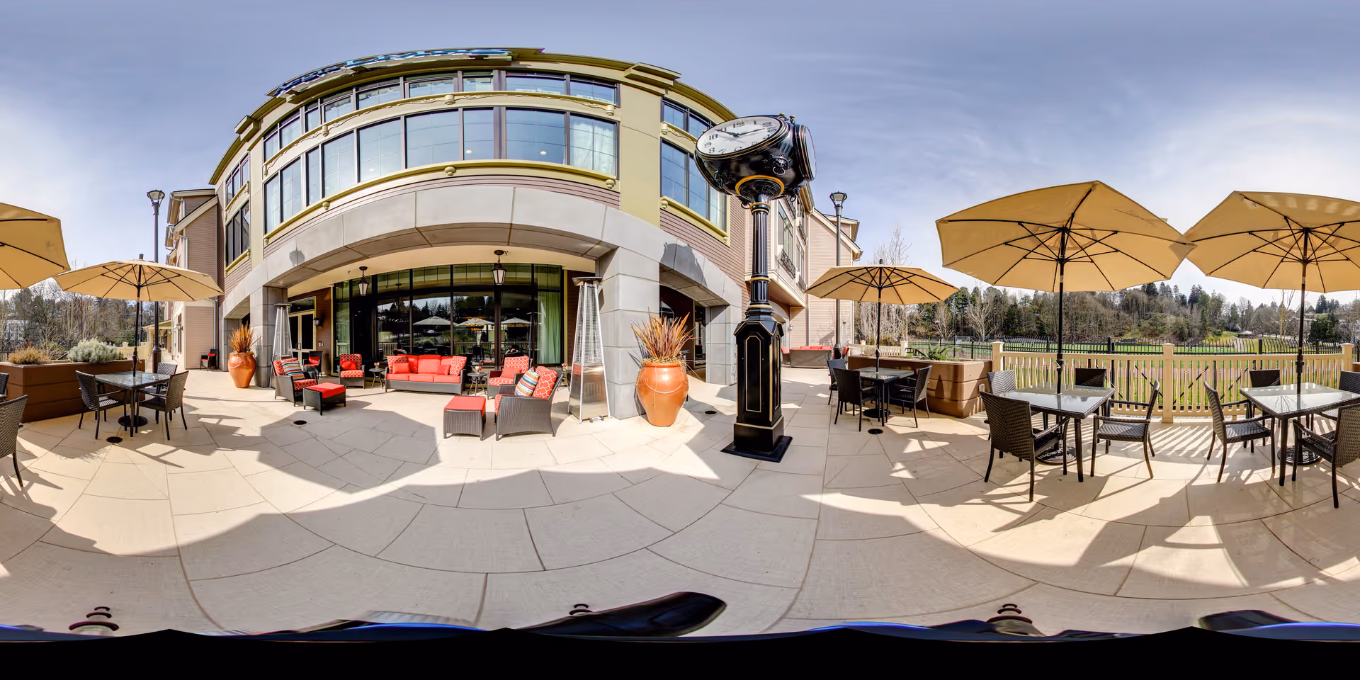 Outdoor patio area at Aegis Living Queen Anne Rodgers Park featuring multiple tables with umbrellas, wicker chairs, and red cushioned seating near the building. A large vintage-style clock stands prominently on the patio, with a scenic view of trees and greenery in the background under a clear sky.