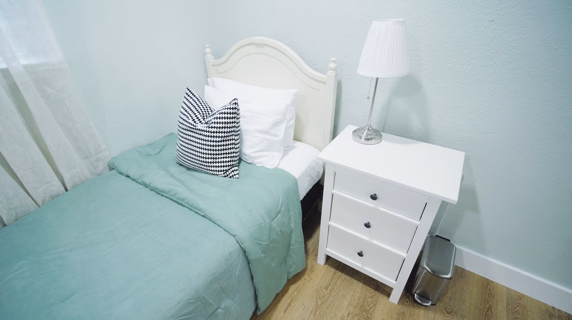 A small bedroom with a single bed featuring a white headboard, white pillows, a black and white patterned throw pillow, and a light green blanket. Next to the bed is a white nightstand with three drawers and a white table lamp. A small silver trash can is on the floor beside the nightstand. The room has light-colored walls and wood flooring.