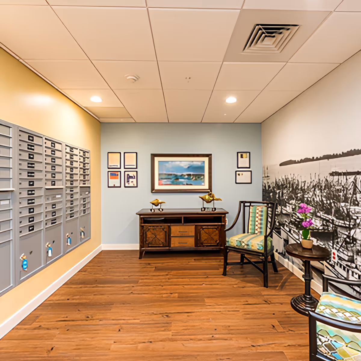 An interior mailroom/lobby with wall mailboxes on the left, wood flooring, a small wooden desk and chairs, framed artwork on the far wall, and a decorative mural on the right.