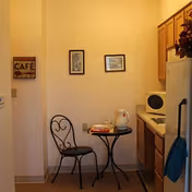 Small kitchenette/dining nook with a round bistro table and chair against a wall, framed pictures, cabinets, microwave, and refrigerator.