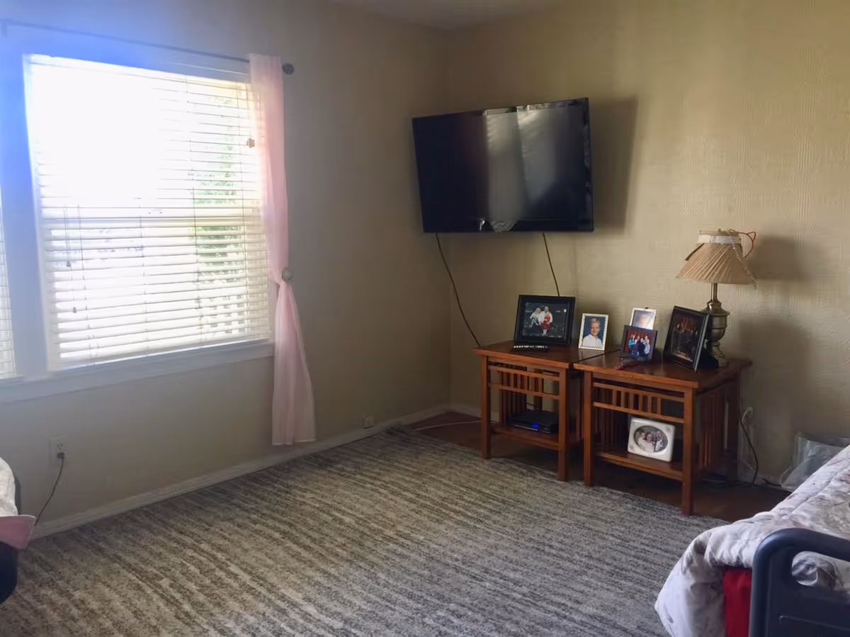 Small living area with a wall-mounted TV, two wooden side tables displaying framed photos and a lamp, a window with blinds and a pink curtain, and a carpeted floor.