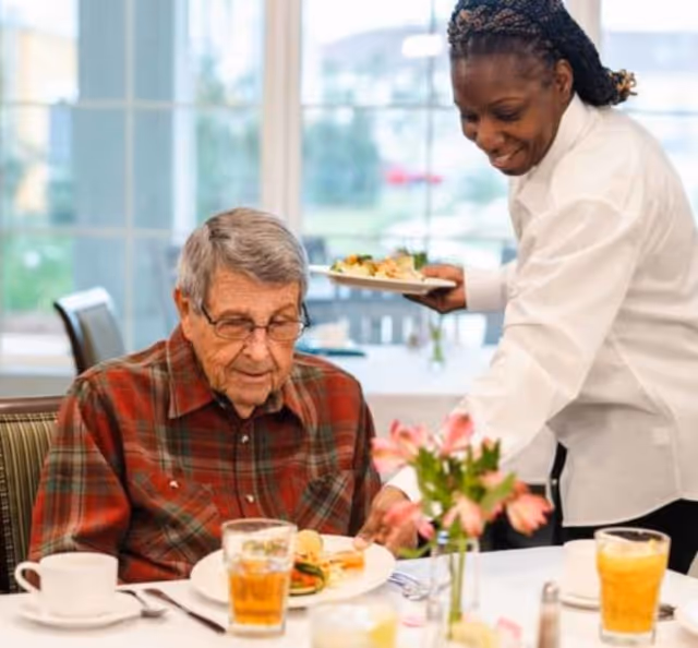 An elderly man wearing glasses and a red plaid shirt is seated at a dining table with a plate of food and a glass of iced tea in front of him. A smiling woman in a white uniform is serving him another plate of food. The setting appears to be a bright dining room with large windows in the background and a small vase of pink flowers on the table.
