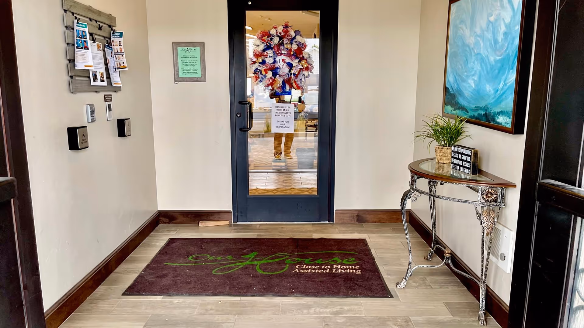 Entrance area of Our House Assisted Living of Tremonton featuring a glass door decorated with a colorful wreath. A brown floor mat with the facility's name and slogan is placed on the tiled floor. To the right, there is a decorative table with a small plant and a framed quote, and a large abstract painting hangs on the wall. On the left wall, there is a bulletin board with flyers and notices.