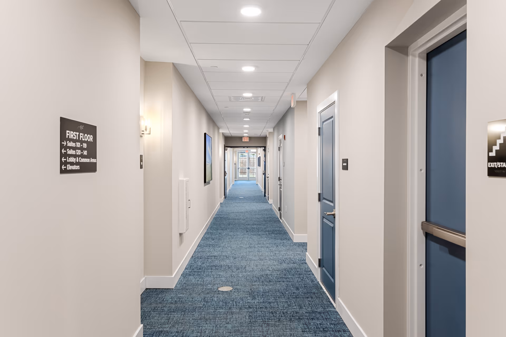 A long, well-lit hallway in a senior living facility with blue carpet and beige walls. There are several blue doors along the right side and a sign on the left wall indicating directions to suites, lobby, common areas, and elevators. The hallway leads to a glass door at the far end.