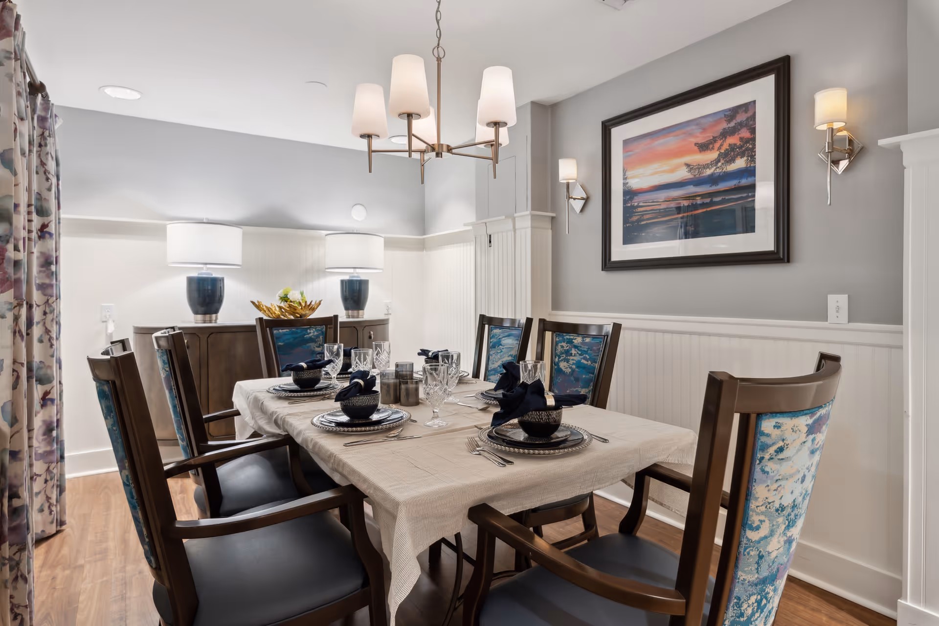 A dining room with a rectangular table covered with a beige tablecloth, set with black bowls, plates, silverware, and glassware. Six wooden chairs with blue patterned upholstery surround the table. The room features a wooden sideboard with two blue lamps and a decorative bowl. A framed landscape painting hangs on the gray wall, and two wall sconces provide additional lighting. A chandelier with five lampshades hangs above the table. The floor is wooden, and floral curtains are partially visible on the left side.