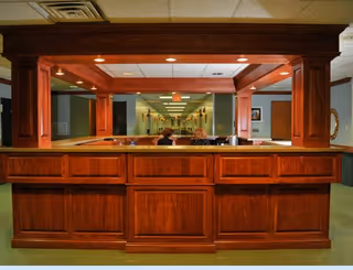 A wooden reception desk with a large rectangular opening and overhead lighting in a senior living facility. Two people are seated behind the desk, and a long hallway with doors and lights extends into the background.