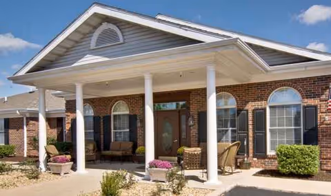 Brick senior living facility front entrance with columns, arched windows, and porch seating, potted plants.