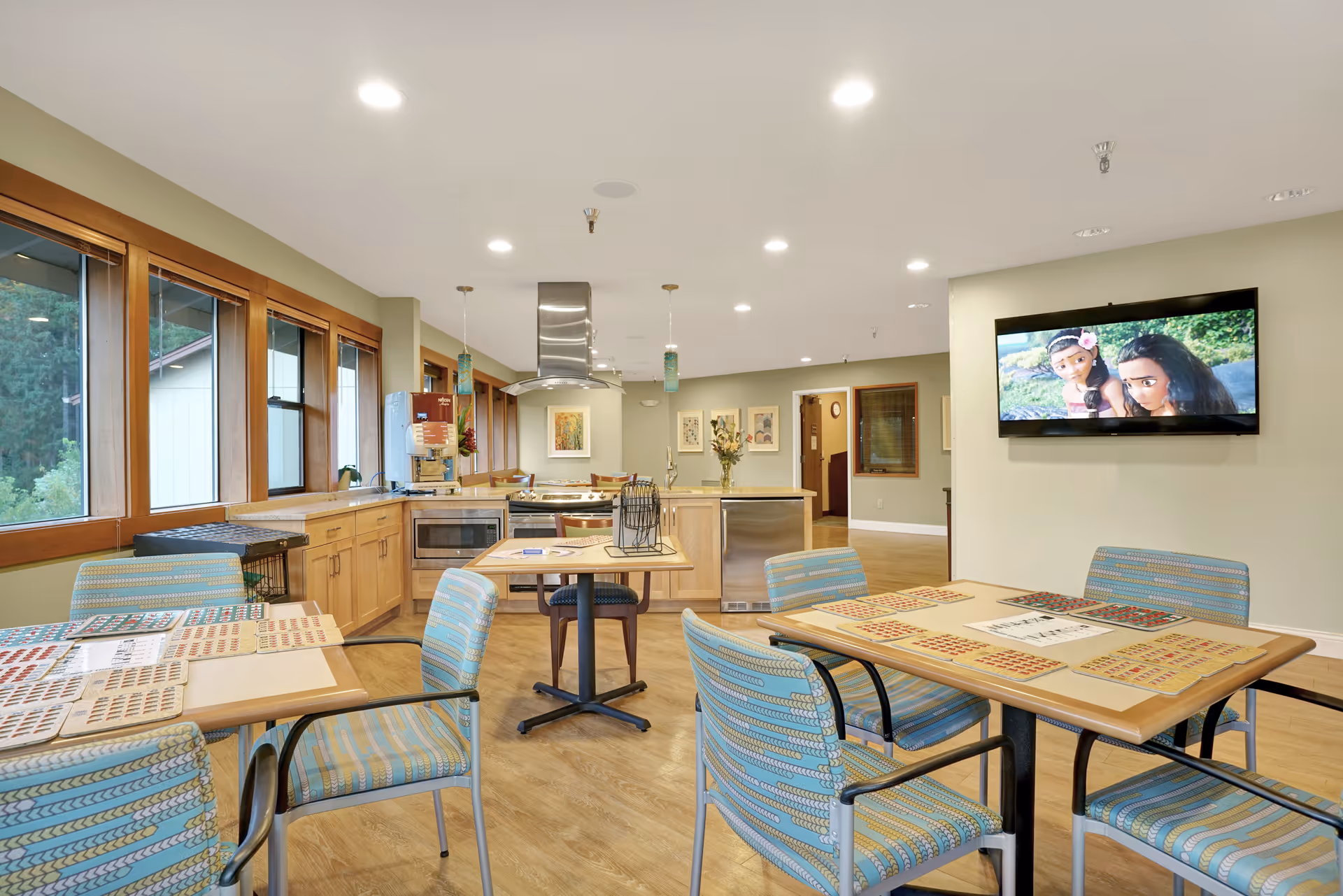 A bright and spacious common area with several tables and chairs arranged for group activities. The tables have bingo cards and markers on them. The room features large windows with wooden frames letting in natural light, a kitchen area with wooden cabinets and stainless steel appliances, and a wall-mounted TV displaying an animated movie.