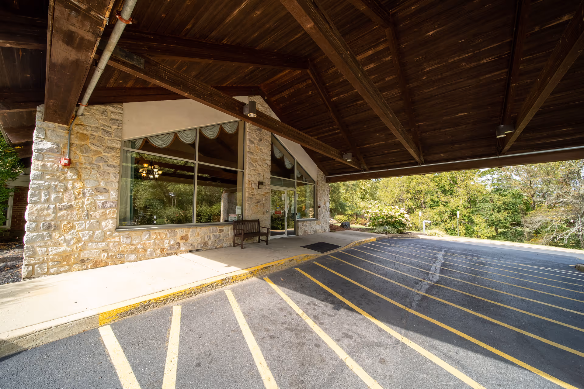 Covered porte-cochere and entrance with stone walls, large windows, a bench, and a striped drop-off driveway.