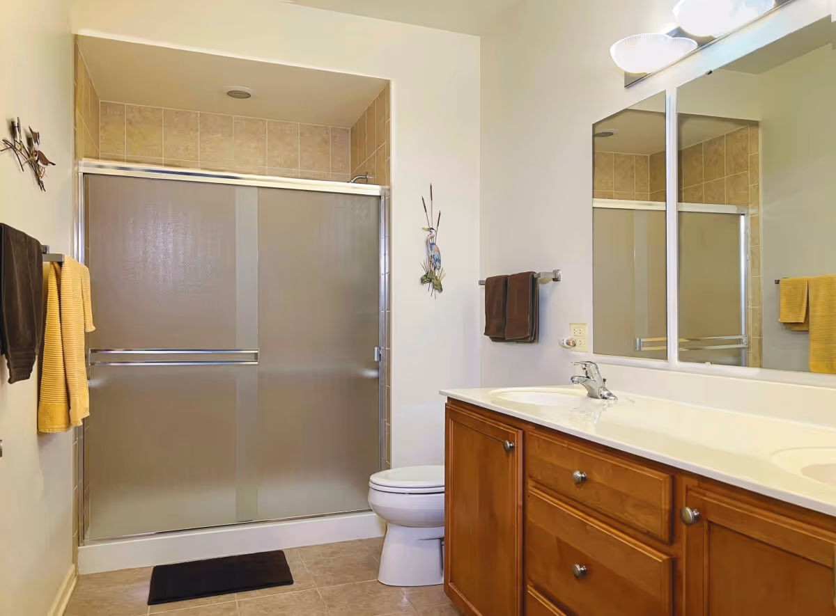 A clean bathroom with a wooden vanity featuring a white countertop and a single sink. Above the vanity is a large mirror with a light fixture. To the left is a toilet and a shower with frosted sliding glass doors and beige tiled walls. Brown and yellow towels hang on towel racks on the walls.