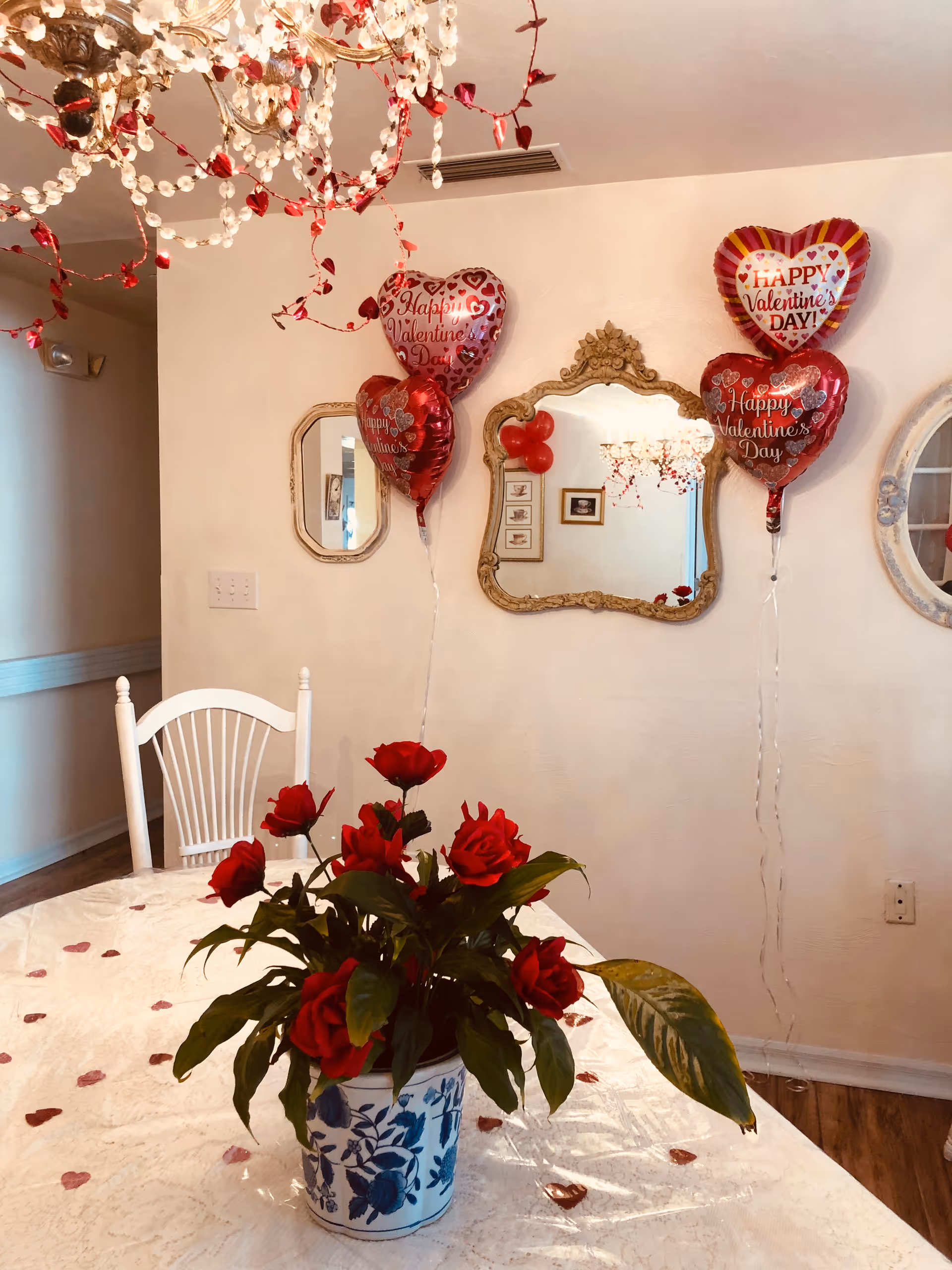 A dining room decorated for Valentine's Day with heart-shaped balloons that say 'Happy Valentine's Day' attached to the wall. A white table with a lace tablecloth is adorned with a potted plant featuring red roses. A white wooden chair is positioned at the table. Three ornate mirrors hang on the wall, and a chandelier with red and white decorations is visible on the ceiling.