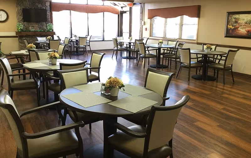 A bright dining room with several tables and chairs arranged neatly. Each table has a small flower arrangement and placemats. The room has large windows with blinds, a wall-mounted TV above a stone fireplace, and a painting on the wall. The floor is wooden, and the overall atmosphere is clean and welcoming.