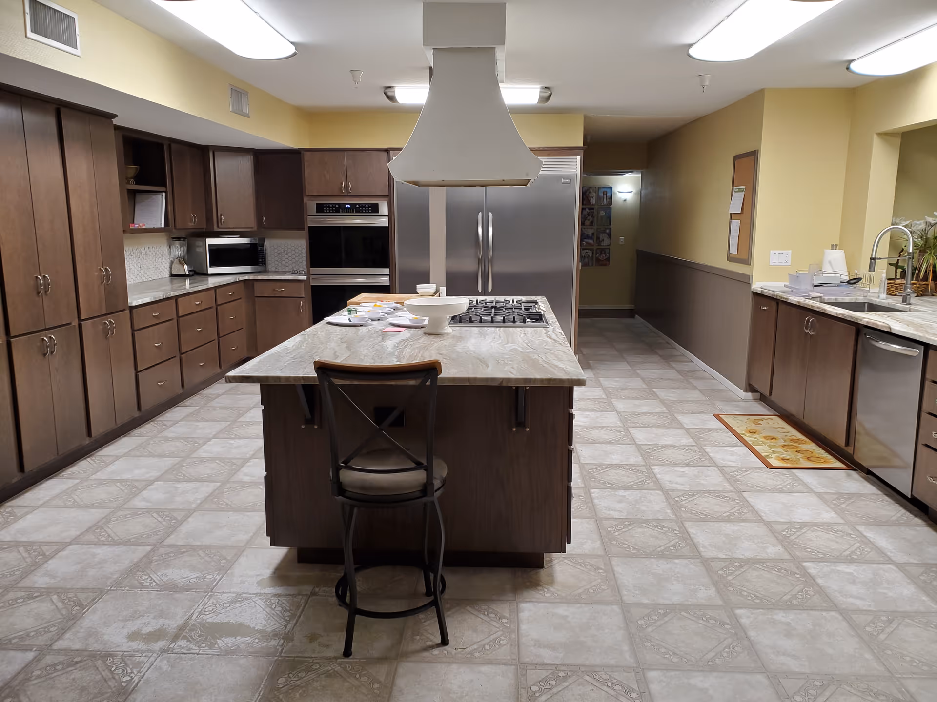 Spacious assisted-living communal kitchen with a central island, stove, refrigerator and surrounding cabinetry.