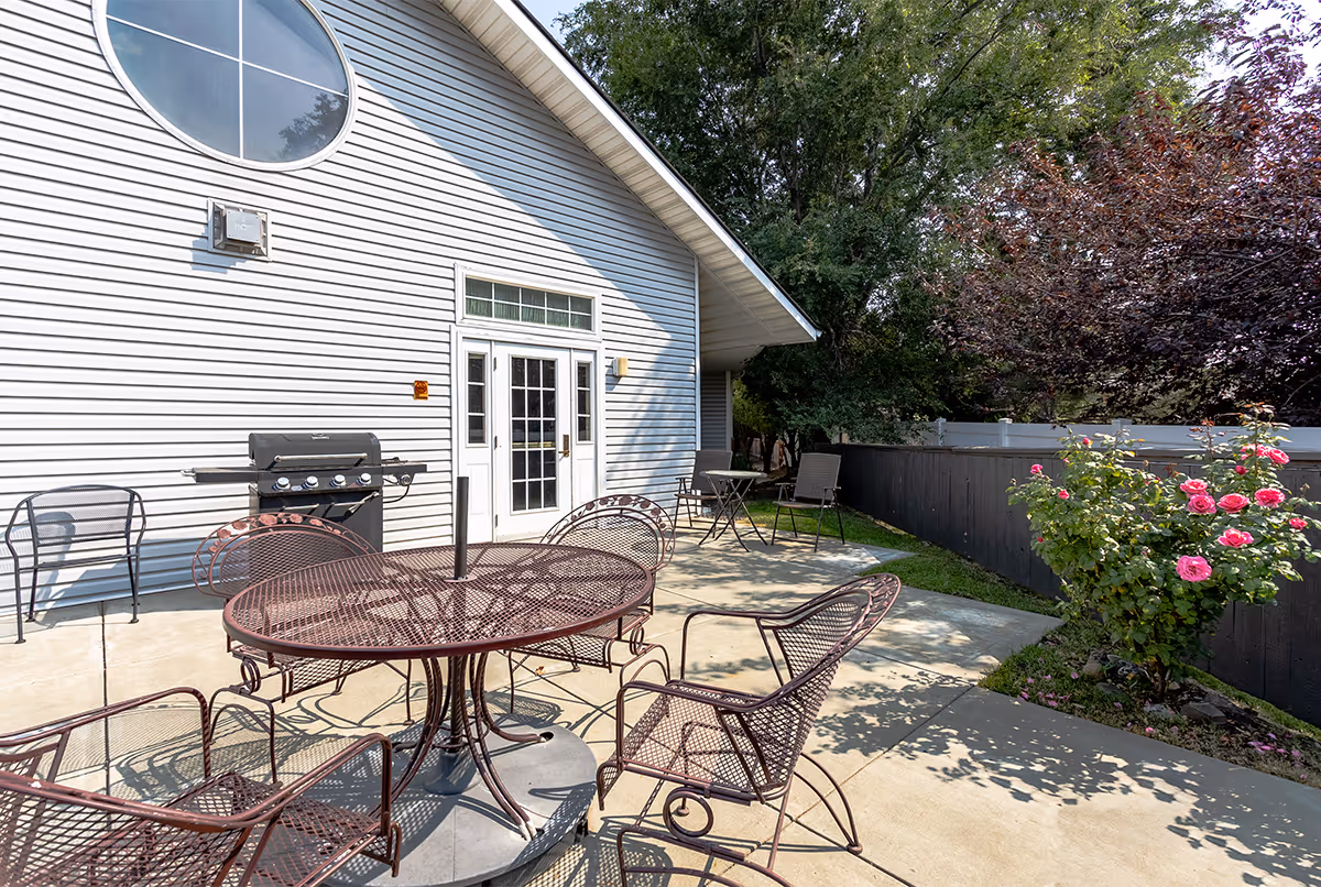 Outdoor patio area with metal table and chairs, a barbecue grill, and a small table with two chairs next to a building with white siding and a round window. There are trees and a flowering bush with pink roses along a black fence.
