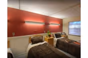 A cozy bedroom with two single beds, each with brown bedding and pillows. Between the beds is a wooden nightstand with a small plant and a framed photo. The walls are painted red with white wainscoting, and there are two modern wall-mounted lights above the beds. A window with blinds is visible on the right side of the room.