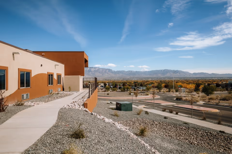 Exterior view of a Southwestern-style assisted living building with a sidewalk, gravel landscaping, and distant mountains under a blue sky.