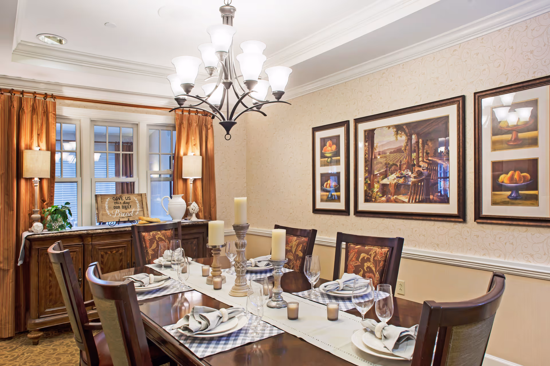 Formal dining room with a set wooden table, candles and place settings, chandelier overhead, framed artwork on the wall and a sideboard beneath curtained windows.
