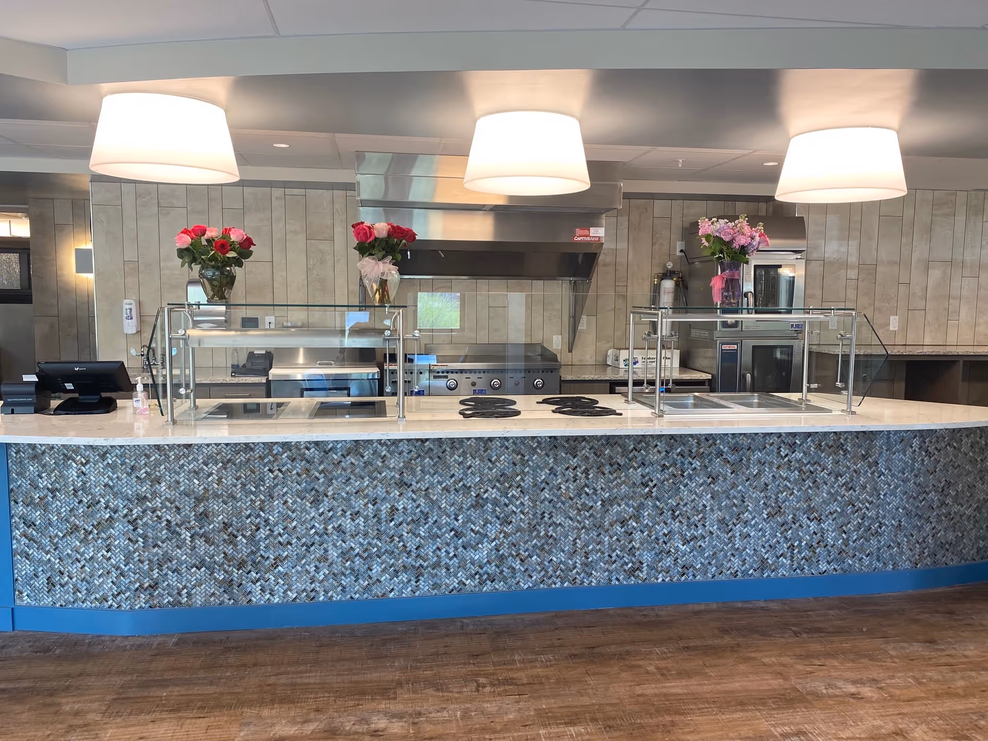 A modern kitchen serving counter with a mosaic tile front and a white countertop. On the counter, there are two glass vases with pink and red flowers. Behind the counter, there is a stainless steel stove, oven, and ventilation hood, along with other kitchen appliances and equipment. Three large ceiling lights hang above the counter, illuminating the area.