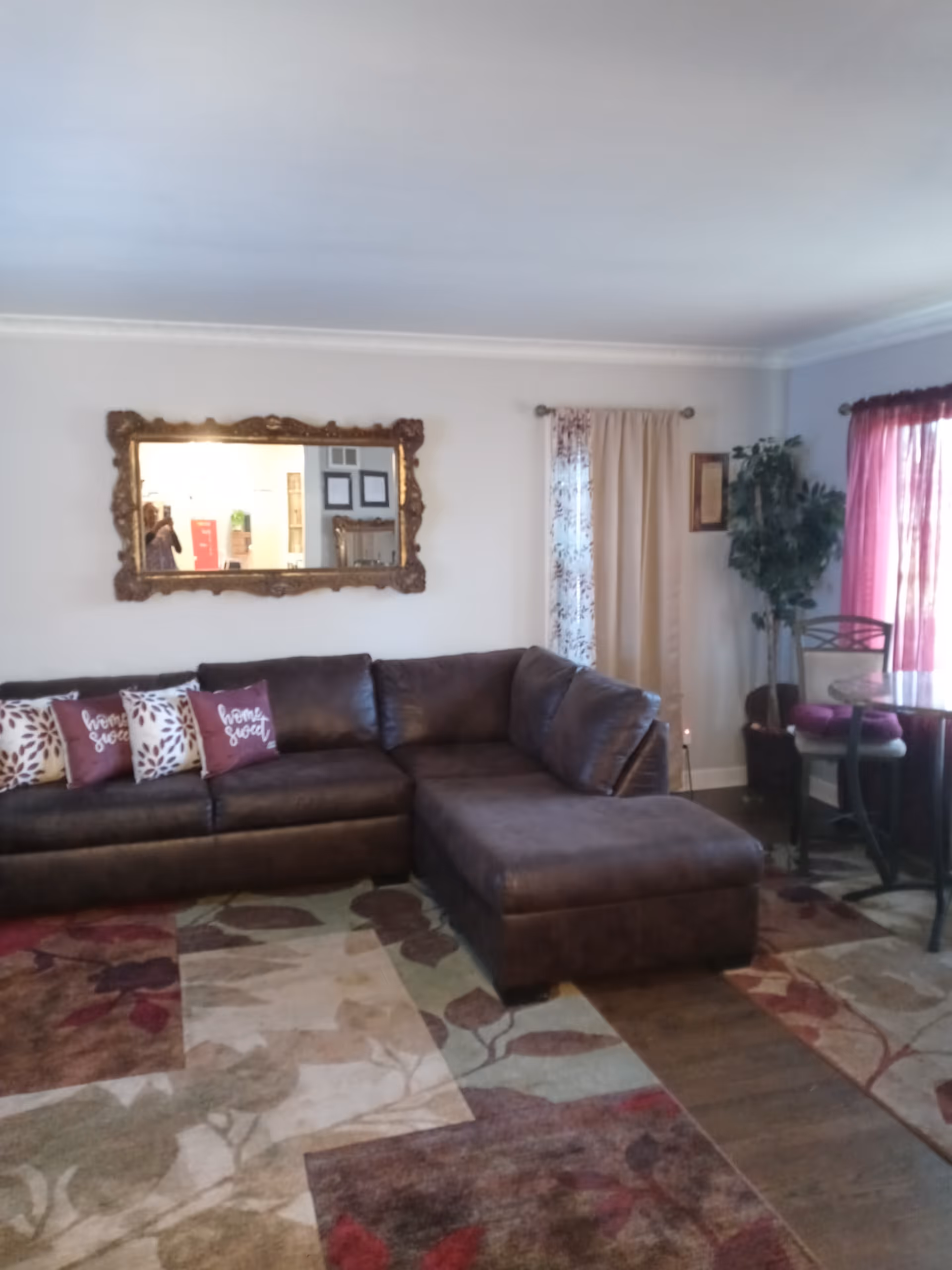 A cozy living room with a large dark brown sectional sofa adorned with decorative pillows, including two with the words 'home sweet'. The room features a patterned area rug with floral designs, a large ornate mirror on the wall, light-colored curtains, a potted plant, and a small round table with chairs near a window with red curtains.