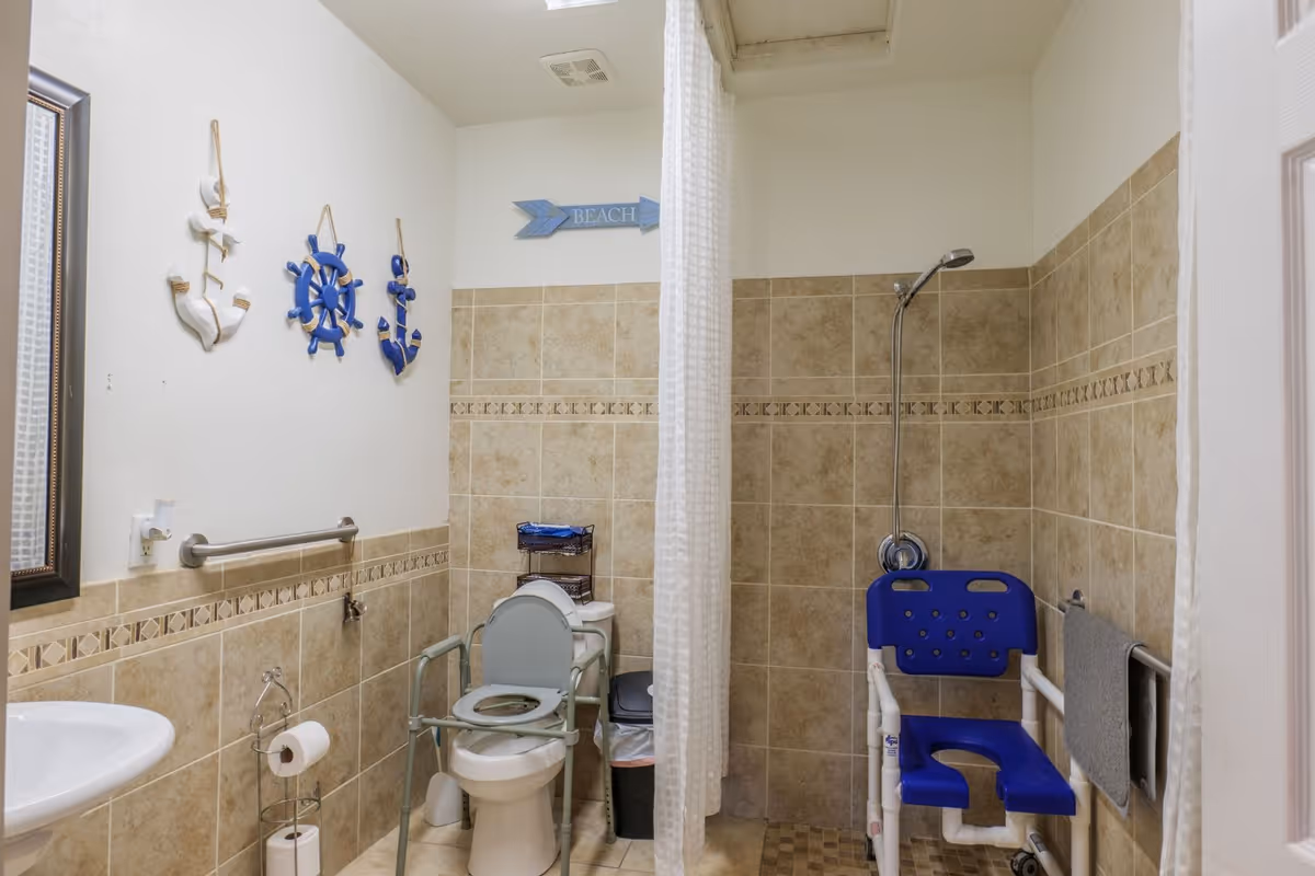 A senior-friendly bathroom with beige tiled walls and floor. The room features a toilet with safety rails and a raised seat, a white sink with a mirror above it, and a shower area with a blue shower chair and a handheld showerhead. There are nautical-themed wall decorations including anchors and a ship wheel, and a sign that says 'BEACH'. A white shower curtain separates the shower area from the rest of the bathroom.
