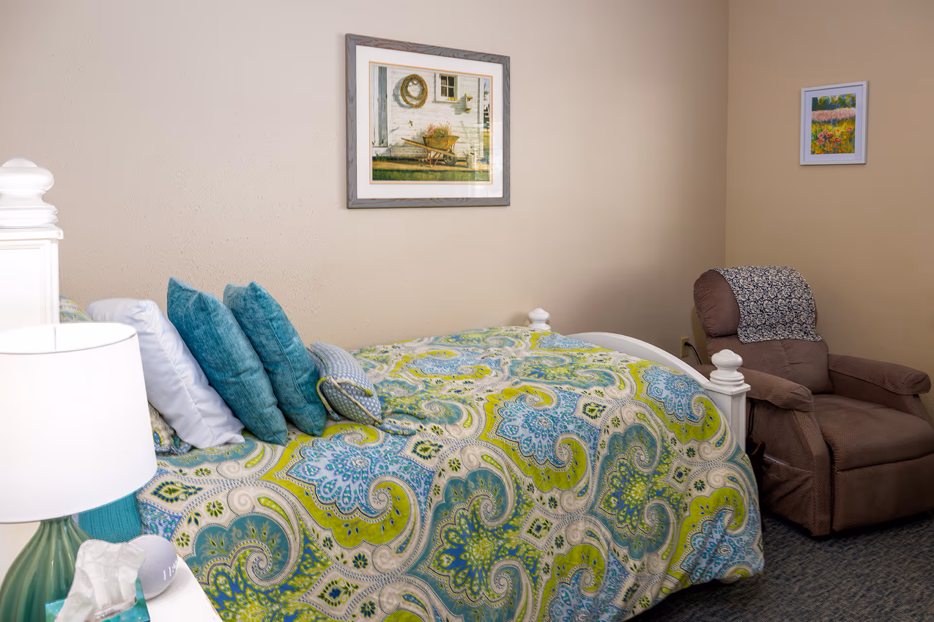 A cozy bedroom in a senior living facility featuring a bed with a colorful paisley-patterned bedspread in shades of green, blue, and white. The bed has multiple pillows in blue and white. Next to the bed is a white nightstand with a green lamp and a box of tissues. On the right side of the room is a brown recliner chair with a patterned cover on the headrest. The walls are beige and decorated with two framed pictures, one above the bed showing a rustic outdoor scene and a smaller floral painting on the adjacent wall.