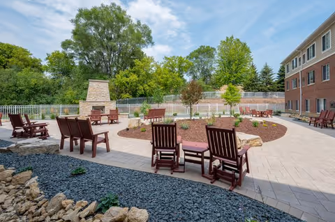 Outdoor patio area at Regent at Burnsville featuring multiple wooden chairs and small tables arranged around a circular paved space with a stone fireplace. The area is surrounded by greenery and trees, with a multi-story brick building visible on the right side under a partly cloudy sky.