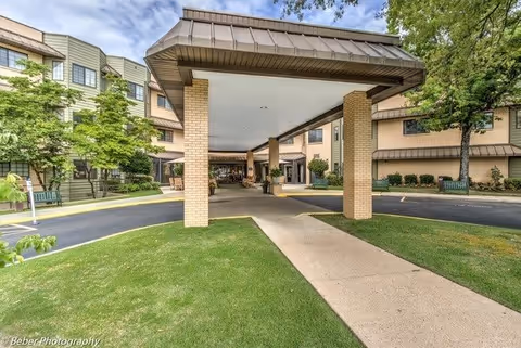 Entrance area of a senior living facility named West Shores, featuring a covered drop-off zone supported by brick columns, surrounded by green lawns, trees, and benches. The multi-story building has a mix of beige and green exterior walls with windows and a brown roof.