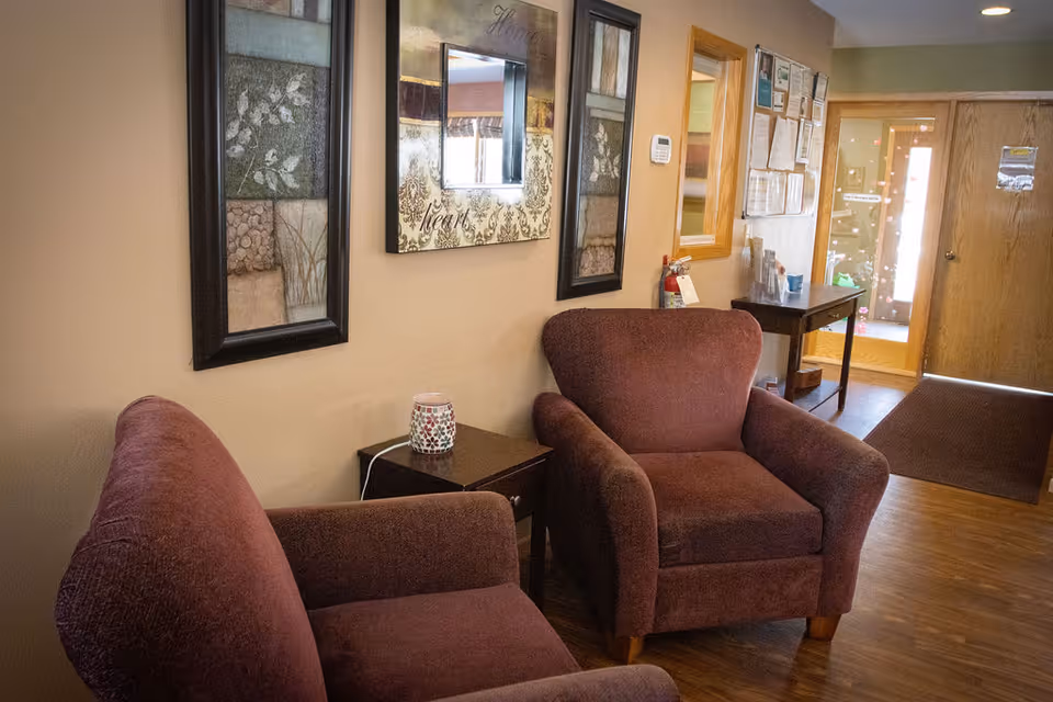A cozy seating area in a hallway with two maroon upholstered armchairs, a small wooden side table with a decorative item on it, and framed artwork and a mirror on the beige wall. The hallway has wooden flooring and leads to a door with glass panels.