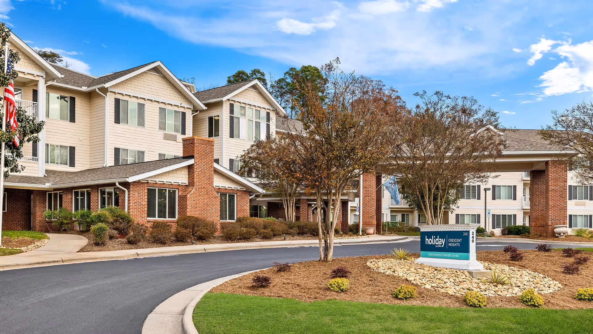 Exterior view of Holiday Crescent Heights senior living facility showing a three-story building with beige siding and brick accents, a covered entrance, landscaped grounds with trees and shrubs, and a sign displaying the facility's name.