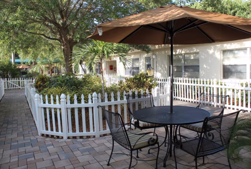 Outdoor patio area with a round metal table and four metal chairs with cushions, shaded by a large brown umbrella. The patio is paved with stone tiles and surrounded by a white picket fence enclosing green plants and trees. A building with multiple windows is visible in the background.