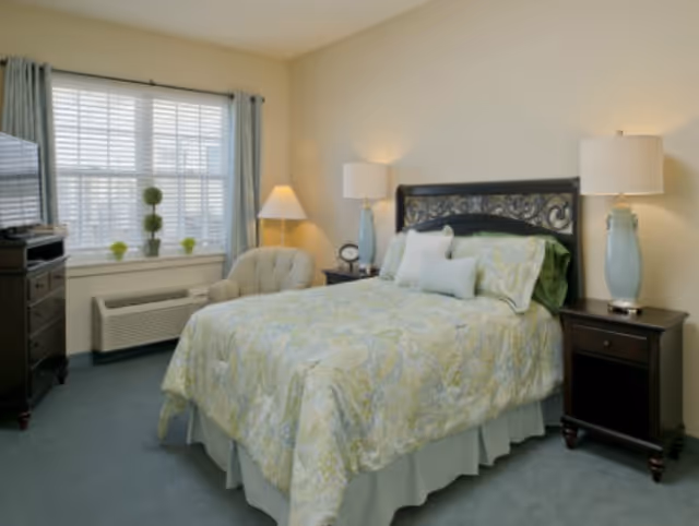 Bright furnished bedroom with a patterned bed, matching nightstands and lamps, an upholstered chair, dresser with TV, and a window with curtains.