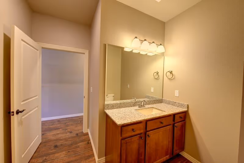 Interior view of a bathroom area in Cedar Springs Independent Living featuring a granite countertop with a built-in sink, wooden cabinets underneath, a large mirror above the sink, and a light fixture with four bulbs. The bathroom door is open, showing a glimpse of an adjacent room with wooden flooring.