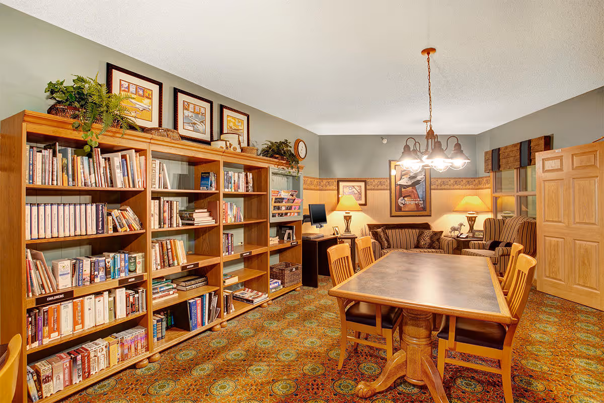 Cozy community room with large bookshelves, a wooden dining table and chairs, and a seating area with lamps.