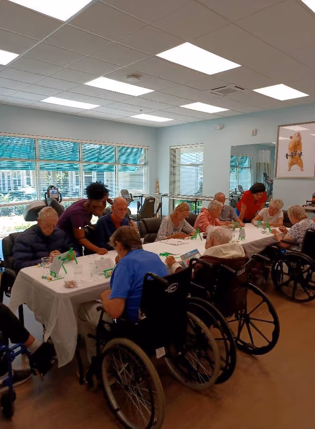 A group of elderly individuals, some in wheelchairs, seated around a long table in a bright room with large windows. Two caregivers are assisting the residents with an activity involving small easels and art supplies. The room has a light blue wall, a large mirror, and a framed picture of a dog lifting weights.